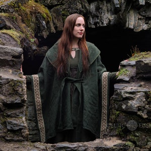 May include: A woman wearing a long, dark green hooded cloak with a brown and white trim stands in a stone doorway. The cloak is tied at the waist with a brown belt. The woman has long brown hair and is looking to the right.