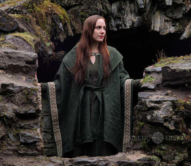 May include: A woman wearing a long, dark green hooded cloak with a brown and white trim stands in a stone doorway. The cloak is tied at the waist with a brown belt. The woman has long brown hair and is looking to the right.