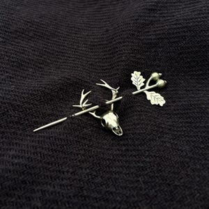 May include: A silver-toned metal brooch set featuring a deer skull and an oak leaf design. The deer skull brooch has a pin on the back. The oak leaf brooch has two leaves and two acorns. The brooches are displayed on a dark gray textured surface.