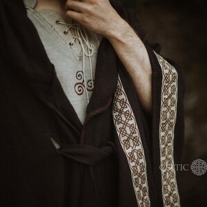 May include: A dark brown robe with a white and brown trim. The robe has a Celtic knot design on the trim. The robe is tied at the waist with a matching sash. The robe is worn over a light gray shirt with a Celtic knot design.