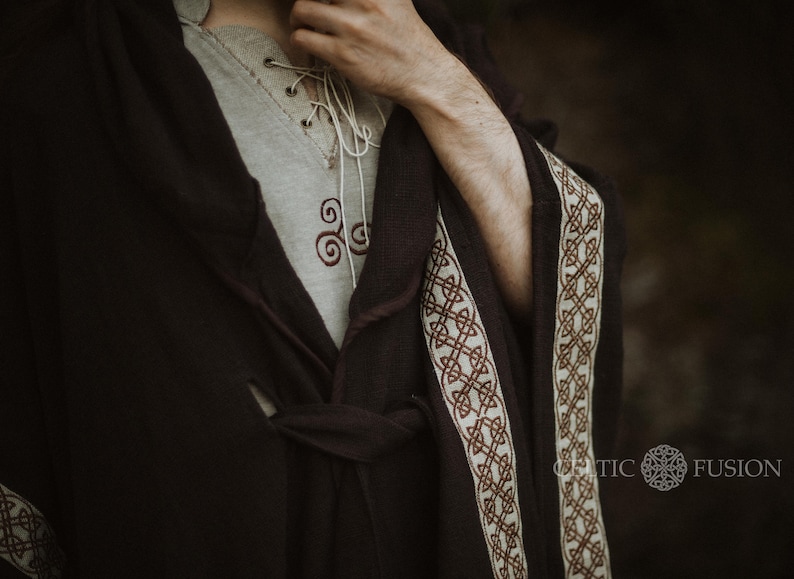 May include: A dark brown robe with a white and brown trim. The robe has a Celtic knot design on the trim. The robe is tied at the waist with a matching sash. The robe is worn over a light gray shirt with a Celtic knot design.