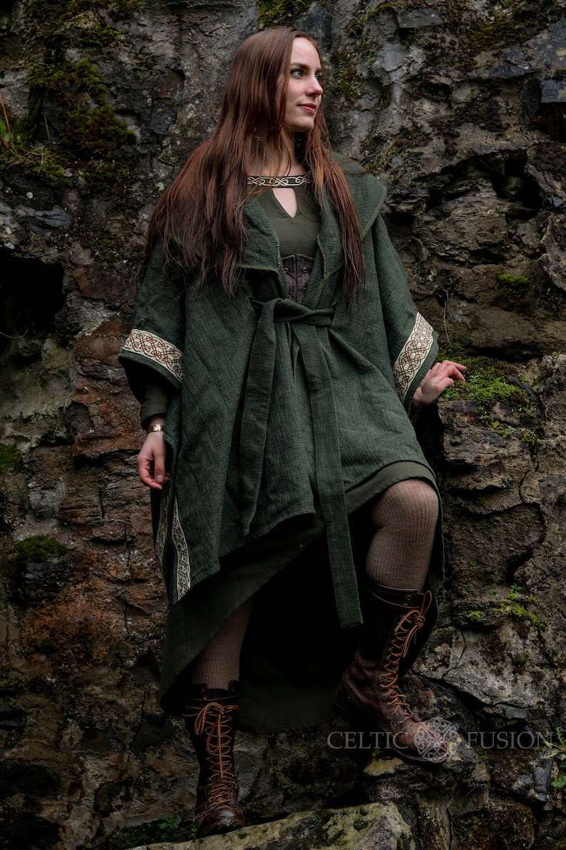May include: A woman wearing a long, dark green hooded cloak with a brown belt and intricate embroidery. She is standing in front of a stone wall with moss growing on it. She is wearing brown leather boots.