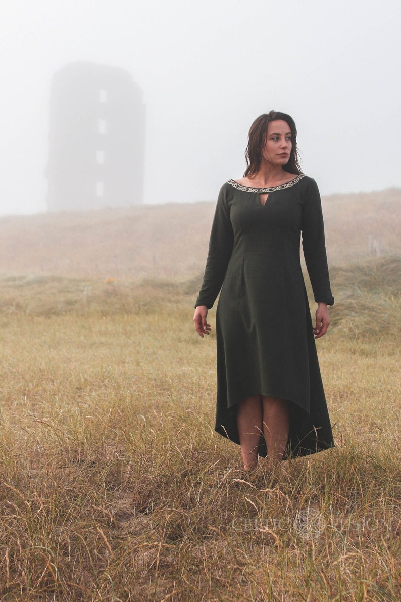 Green Celtic dress with Celtic knot embroidery, worn as an alternative wedding guest dress with high low skirt in a foggy landscape.