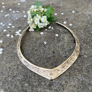 May include: A gold-toned, V-shaped choker necklace with a floral design. The necklace is lying on a grey rock with white flower petals scattered around it.