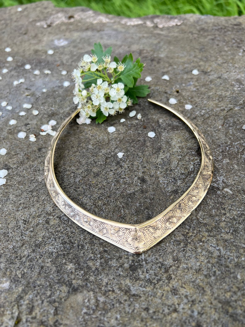 May include: A gold-toned, V-shaped choker necklace with a floral design. The necklace is lying on a grey rock with white flower petals scattered around it.