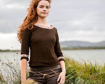 May include: A brown, long-sleeved shirt with a Celtic knot design on the neckline and cuffs. The shirt has a fringe trim at the bottom.