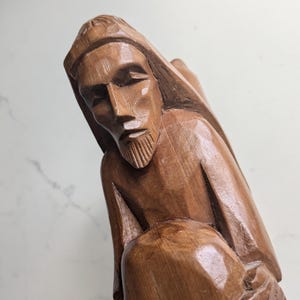 May include: A carved wooden sculpture of a figure with a beard and hooded garment. The sculpture is a medium brown colour and has a detailed carving of the face and clothing. The figure appears to be in a seated position.