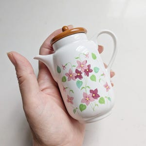 Peut inclure: Une petite théière en céramique blanche ornée d'un motif floral composé de fleurs roses et violet foncé avec des feuilles vertes. La théière présente un corps blanc lisse, une anse courbée, un bec court et un couvercle marron brillant.