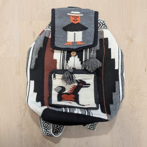 May include: A woven fabric backpack featuring a geometric pattern in black, white, gray, and brown. The top flap displays a woven figure wearing a hat, while the front pocket features a woven animal graphic. It includes gray tassels and a wooden button.