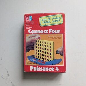 Może przedstawiać: Czerwone pudełko gry podróżnej Connect Four firmy Milton Bradley z żółtą pionową kratką oraz czerwonymi i czarnymi żetonami. Pudełko zawiera napisy Connect Four, Puissance 4 oraz Jeux de voyage, wskazujące na grę dla dwóch osób.