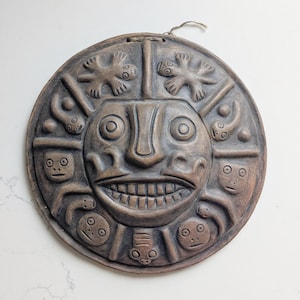 May include: A circular brown clay wall plaque featuring a central face with a wide, toothy grin and large circular eyes. The border is decorated with relief carvings of various small figures, including lizard-like creatures and stylised faces, with a string loop at the top.