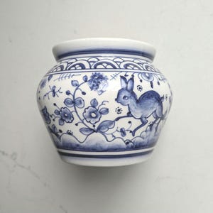 May include: A white ceramic planter featuring a hand-painted blue design of a rabbit surrounded by floral motifs and vines. The pot has a rounded shape with a wide rim and decorative scalloped borders at the top and bottom edges in blue.