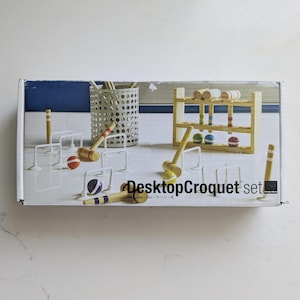Op de afbeelding: Een Desktop Croquet set in een rechthoekige doos met houten hamers met gekleurde strepen, kleine houten ballen, witte draadpoortjes en een houten opbergrek. De doos bevat de tekst Desktop Croquet set met een klein logo aan de rechterkant.