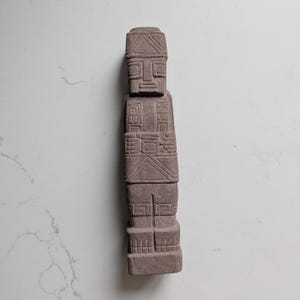 Puede incluir: Una escultura de piedra de color marrón claro, alta y rectangular, con patrones geométricos tallados y rasgos faciales estilizados. La figura presenta una cabeza plana, ojos cuadrados y un torso robusto con grabados lineales.