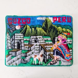 May include: A rectangular, textured textile art piece featuring the words "CUZCO PERU" in red letters. The artwork depicts a colorful scene with mountains, ancient stone structures, a person playing a flute, and a llama.