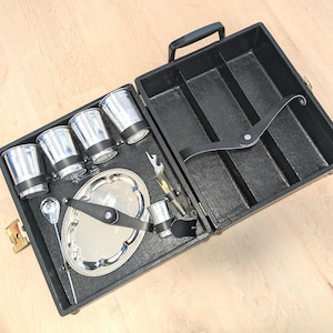 Op de afbeelding: Een zwarte rechthoekige draagkoffer met handvat, met daarin vier zilverkleurige metalen bekers met zwarte banden, een klein ovaal zilveren dienblad, een zilveren lepel met lange steel en een metalen flesopener, vastgezet met zwarte riemen.