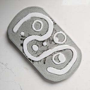 May include: A rectangular ceramic tray with rounded corners, featuring a speckled grey base glaze. The surface is decorated with a raised, glossy white linear design that forms an abstract, continuous path with several circular accents.