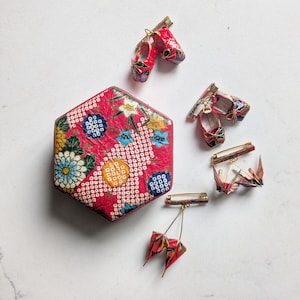 Op de afbeelding: Een zeshoekige doos met een rood bloemenpatroon en drie goudkleurige metalen broches. Twee broches bevatten miniatuurschoenen met een rood patroon en één broche bevat twee rode papieren kraanvogelornamenten die aan een metalen staaf hangen.