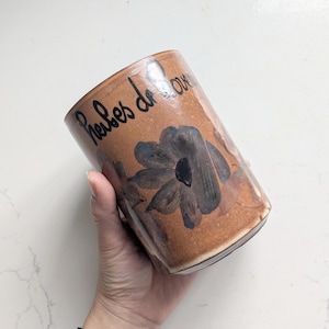 Pode incluir: Um recipiente cilíndrico de cerâmica castanho-claro com acabamento brilhante, apresentando um desenho de flor pintado à mão em azul escuro e o texto em cursivo preto "Herbes de Provence" escrito na parte superior exterior.