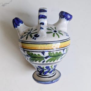 Peut inclure: Un récipient en céramique à base blanche, orné de motifs floraux peints à la main en bleu et vert, d'une bande horizontale jaune et de deux becs. L'objet possède une anse centrale et une base piédouche décorée de feuilles bleues et vertes.