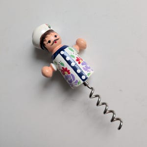 May include: A corkscrew featuring a hand-painted wooden handle shaped like a chef wearing a white hat. The chef figure has a moustache and a white coat decorated with colourful red and purple floral patterns, blue vertical stripes, and white dots.