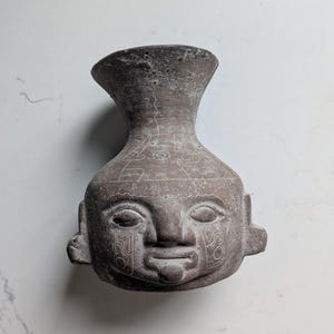 May include: A gray, weathered ceramic vessel shaped like a human head with a flared neck. The surface features etched, linear geometric patterns across the forehead and cheeks, with stylized eyes, a nose, and a mouth carved into the front.
