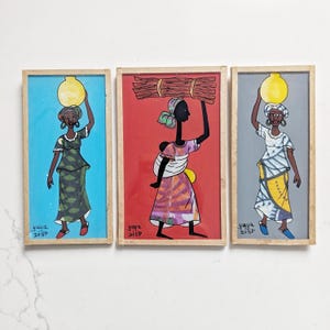 May include: Three rectangular framed paintings by Yaya Diop. The left shows a person in a green patterned dress carrying a yellow pot on their head against a blue background. The center shows a person carrying wood on a red background. The right shows a person in a patterned dress with a yellow pot on a gray background.