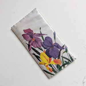 May include: A rectangular white fabric case featuring a botanical print of two large purple iris flowers and one smaller yellow iris flower with green stems and leaves. The case has stitched edges and a smooth, satin-like finish on the surface.