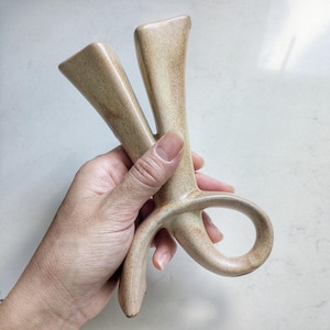 May include: A beige ceramic object with a speckled texture, featuring two upward-pointing prongs and a curved loop at the base. The piece has a smooth, matte finish and an abstract, sculptural form with organic, flowing lines throughout its structure.