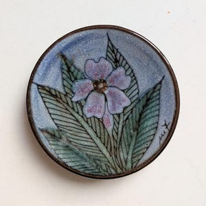 May include: A small, round dish with a floral design. The dish features a light blue background with a purple flower and green leaves. The rim is a dark brown color. The floral design is hand-painted.