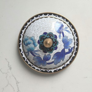 May include: A white cloisonné lid with a blue and green floral design. The lid has a black rim and a gold knob.