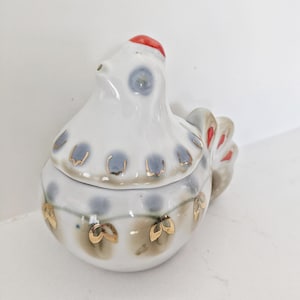 May include: A white ceramic chicken-shaped lidded container featuring a red comb, blue-grey painted accents, and gold-coloured leaf-like details along the body. The lid is shaped like the head and neck of a chicken, resting on a rounded base.