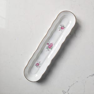 Peut inclure: Un long plat de service en porcelaine blanche avec un bord festonné et une bordure dorée. Le plat est décoré de grappes de roses roses et de feuilles vertes. Une pièce décorative pour servir des amuse-gueules ou de petites friandises.
