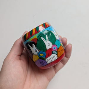 Può includere: Un piccolo contenitore cilindrico in legno con un disegno dipinto a mano di conigli bianchi tra fogliame verde, un sole giallo e forme astratte colorate. La base e il bordo sono dipinti in rosso tinta unita con una finitura lucida.