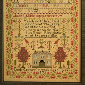 May include: Framed cross-stitch sampler with a cream background. The design includes a house, trees, birds, and floral borders in red, green, and blue. The text includes the alphabet, numbers, and a poem. The sampler is dated 1872.