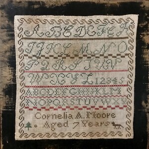 May include: A sampler with a white background and brown, red, and green embroidery. The sampler includes the alphabet, numbers, and the text "Cornelia A. Moore Aged 7 Years".