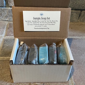 May include: A sample soap set in a cardboard box. The set includes five individually wrapped soaps in various colors, including gray, blue, and black. A white card inside the box lists the ingredients and states the soaps are handmade using the cold process method.