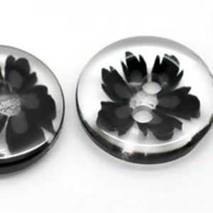 May include: Two clear, round buttons with a black floral design encased within. Each button has two holes for sewing. The buttons are set against a white background, highlighting the contrast between the clear resin and the black floral pattern.