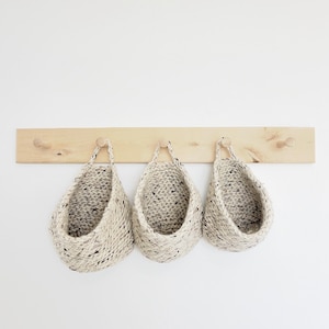 May include: Three beige knitted hanging baskets with black flecks are displayed on a wooden rack with five knobs. The baskets are hanging from the knobs.