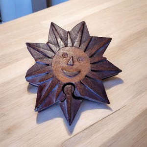 May include: A wooden puzzle box shaped like a sun with a smiling face carved into the front. The box is made of dark wood with a lighter brown sun.