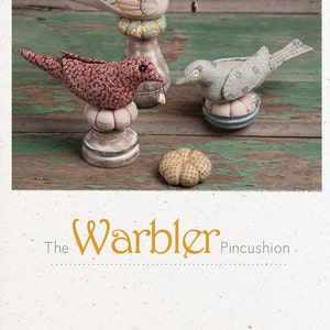 May include: Three bird-shaped pincushions, one beige, one red, and one blue, with a small pumpkin-shaped pincushion. The pincushions are on a wooden surface. The text "The Warbler Pincushion" is below the pincushions.