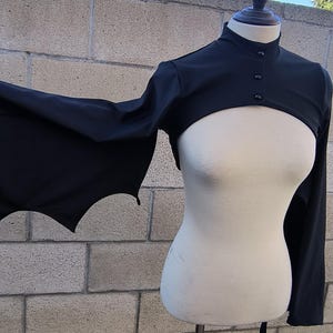 Könnte beinhalten: Ein schwarzer Bolero mit langen Ärmeln, die mit verlängerten, spitz zulaufenden Stoffbahnen im Fledermausflügel-Stil versehen sind. Das Kleidungsstück hat einen hohen Stehkragen und wird vorne mit drei kleinen, runden schwarzen Knöpfen geschlossen.