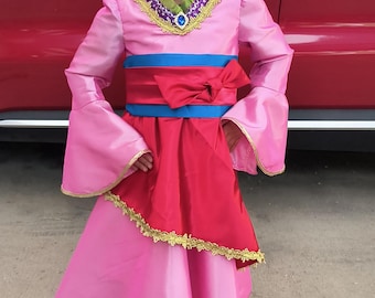 May include: A pink long-sleeved dress with flared sleeves, a green ruffled collar, and a gold-trimmed neckline featuring a blue faux gem. The dress includes a wide blue waistband with a red sash and a red skirt overlay with gold trim.