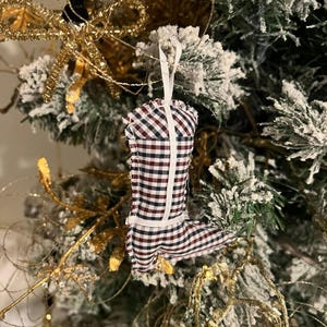 Könnte beinhalten: Ein Weihnachtsbaumschmuck aus Stoff in Form eines Cowboy-Stiefels. Der Stiefel ist aus einem rot-weiß-marineblau karierten Stoff mit weißer Bordüre und einem weißen Band zum Aufhängen gefertigt. Der Schmuck hängt an einem geschmückten Weihnachtsbaum.