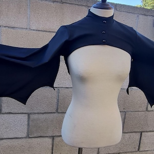 May include: A black bat wing shrug on a mannequin. The shrug has a high collar and three buttons. The wings extend outwards with a scalloped edge, creating a dramatic silhouette. The garment is made of a dark, opaque fabric, perfect for a costume.