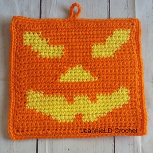 May include: Orange and yellow crocheted pot holder with a jack-o'-lantern face design. The pot holder has a loop for hanging. "JeannieLD Crochet" is written in the bottom right corner.
