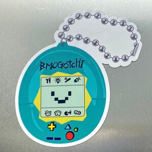May include: A teal-colored BMOgotchi sticker with a pixelated face and a screen displaying icons. The sticker has a chain of silver beads attached to the top. The text "BMOgotchi" is written in blue.