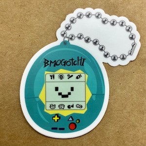 May include: A teal and white sticker featuring a pixelated digital pet design. The sticker has the text "BMOGOTCHI" and a small screen with icons and a smiling face. A chain of silver beads is attached to the top.