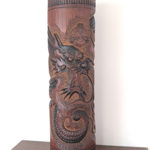 May include: A cylindrical brown wooden vessel featuring intricate relief carvings of a dragon with scales and whiskers. The top and bottom edges are decorated with a repeating vertical notch pattern, and a circular emblem is visible above the dragon.