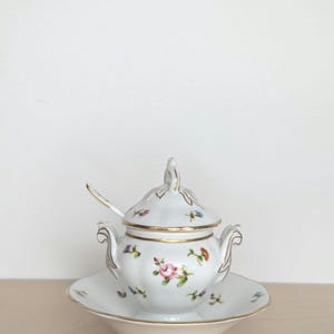 May include: A white porcelain sugar bowl with a lid and a matching saucer. The bowl and saucer are decorated with a floral pattern in shades of pink, blue, and yellow. The bowl has a gold trim around the rim.